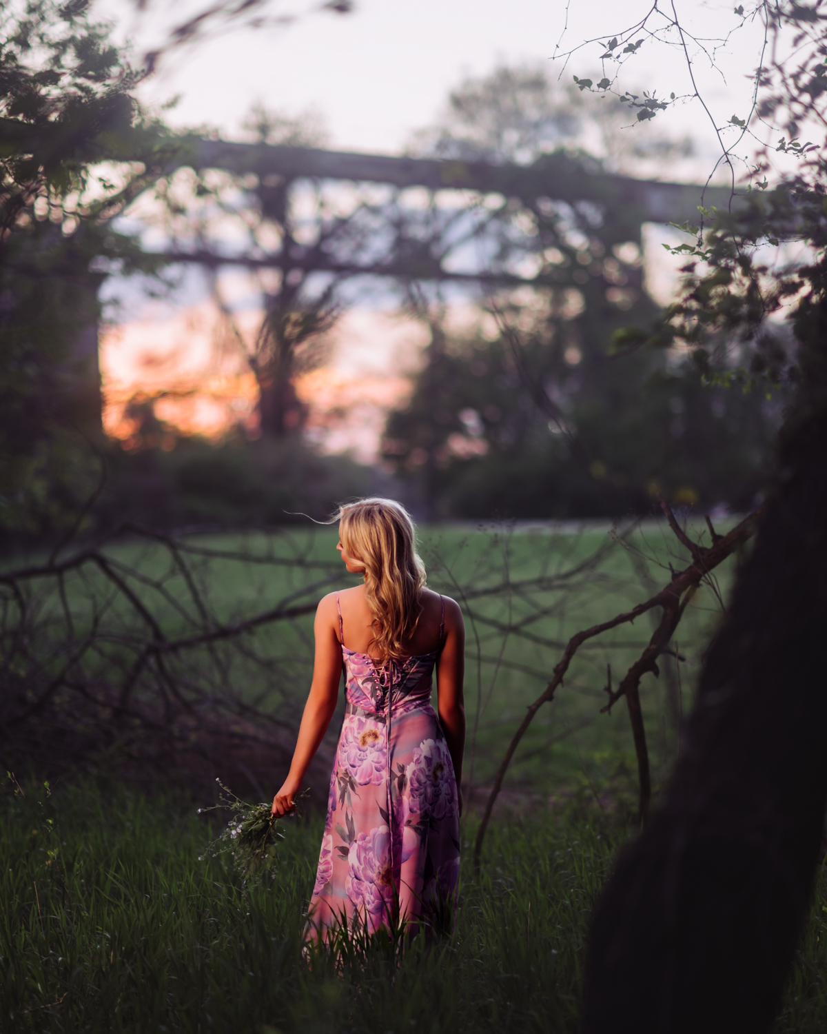 A high school senior in a lavender floral dress stands with her back to the camera in tall grass at Shelby Bottoms, a handful of wildflowers at her side, as a pink and orange sunset glows behind the railroad bridge in the distance. A quiet, dreamy Nashville senior portrait by Cate Connery Bury Photography, captured as she looks toward everything ahead.” A high school senior in a lavender floral dress stands with her back to the camera in tall grass at Shelby Bottoms, a handful of wildflowers at her side, as a pink and orange sunset glows behind the railroad bridge in the distance. A quiet, dreamy Nashville senior portrait by Cate Connery Bury Photography, captured as she looks toward everything ahead.”