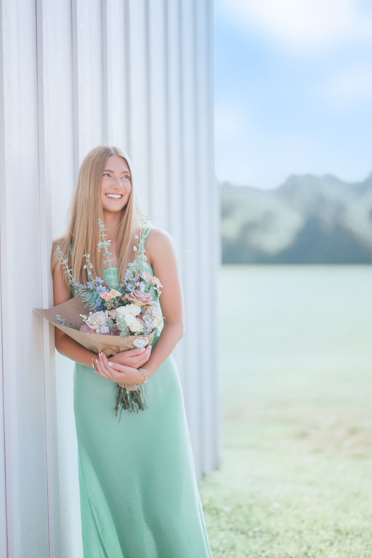 A high school senior in a flowing sage green dress leans against a white board-and-batten wall at Smith Park, laughing as she glances away, a hand-tied bouquet of dusty roses, blue delphinium, and eucalyptus wrapped in kraft paper held close. A soft, airy Brentwood senior portrait by Cate Connery Bury Photography, full of easy joy and summer light. A high school senior in a flowing sage green dress leans against a white board-and-batten wall at Smith Park, laughing as she glances away, a hand-tied bouquet of dusty roses, blue delphinium, and eucalyptus wrapped in kraft paper held close. A soft, airy Brentwood senior portrait by Cate Connery Bury Photography, full of easy joy and summer light.