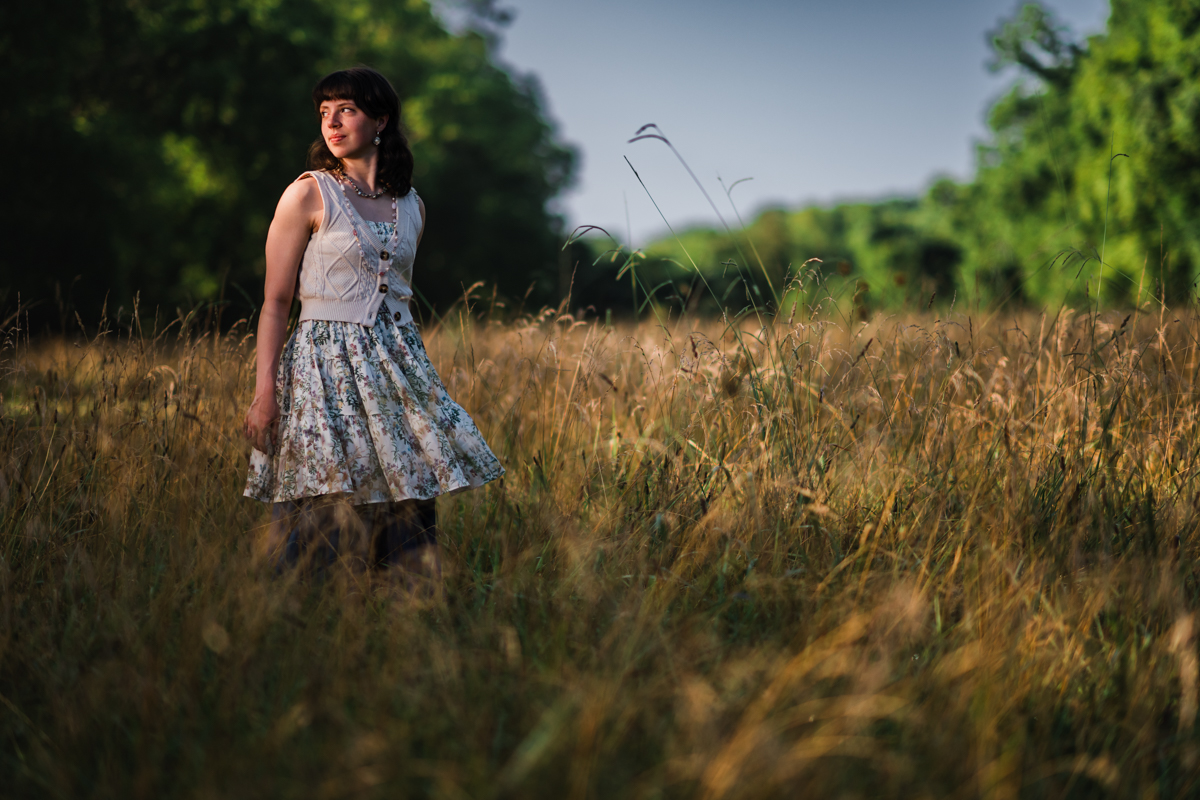 Nashville high school senior in a floral skirt and cream knit vest stands in tall golden grass, glancing over her shoulder as warm light finds her face.