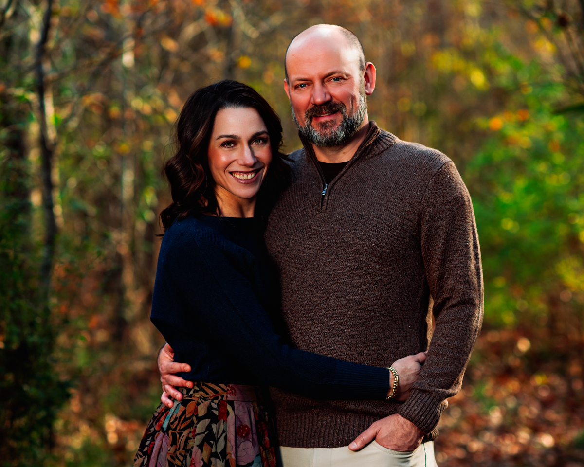 A husband and wife lean into each other on a golden autumn trail, her arm wrapped around his waist and his arm holding her close, both smiling with the easy warmth of years together. Fall couples portrait by Nashville family photographer Cate Connery Bury.