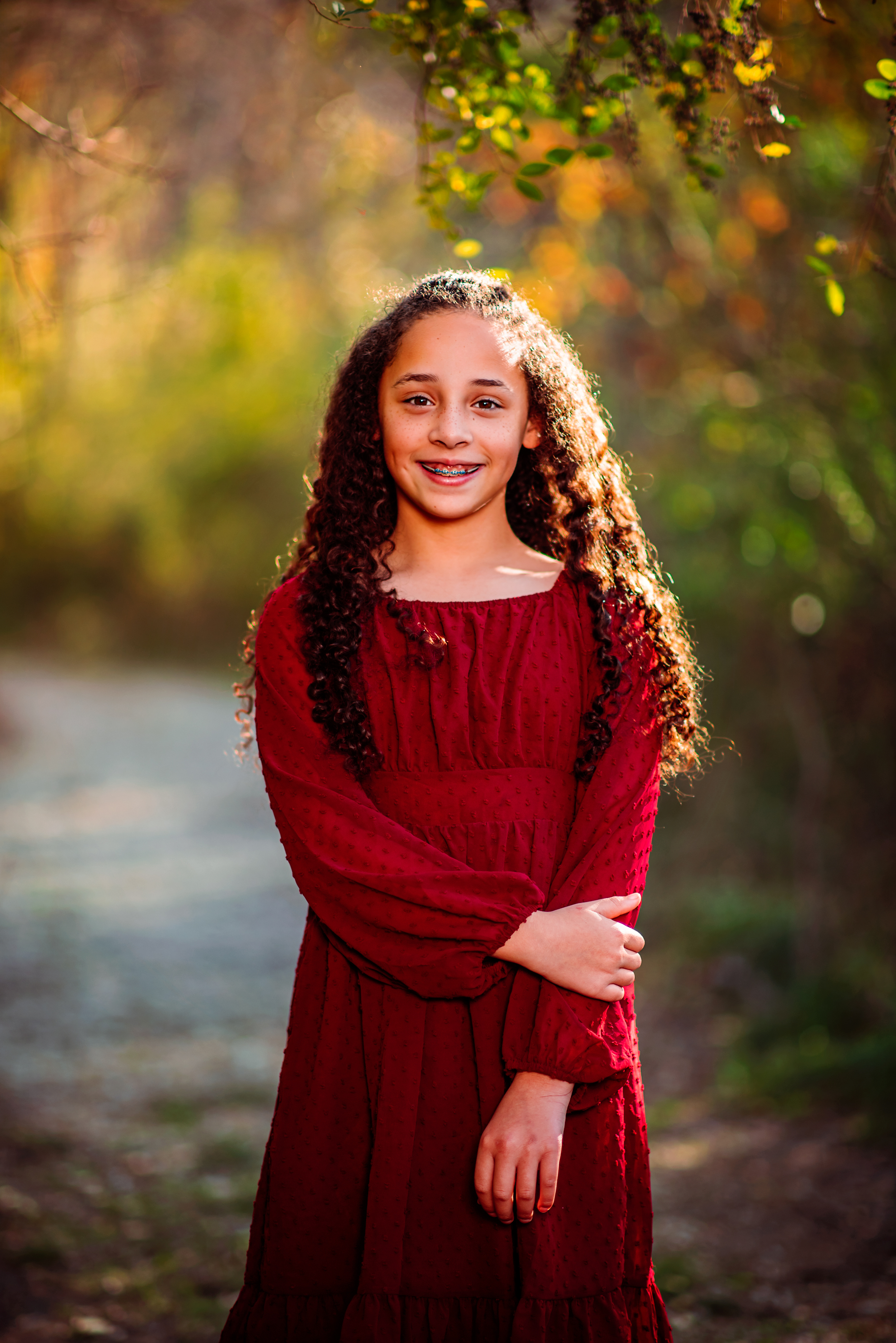 A young girl in a deep red dress smiles shyly, one hand resting on her arm, as late-afternoon sun lights up her long curls on a golden autumn trail. Fall child portrait by Nashville family photographer Cate Connery Bury.
