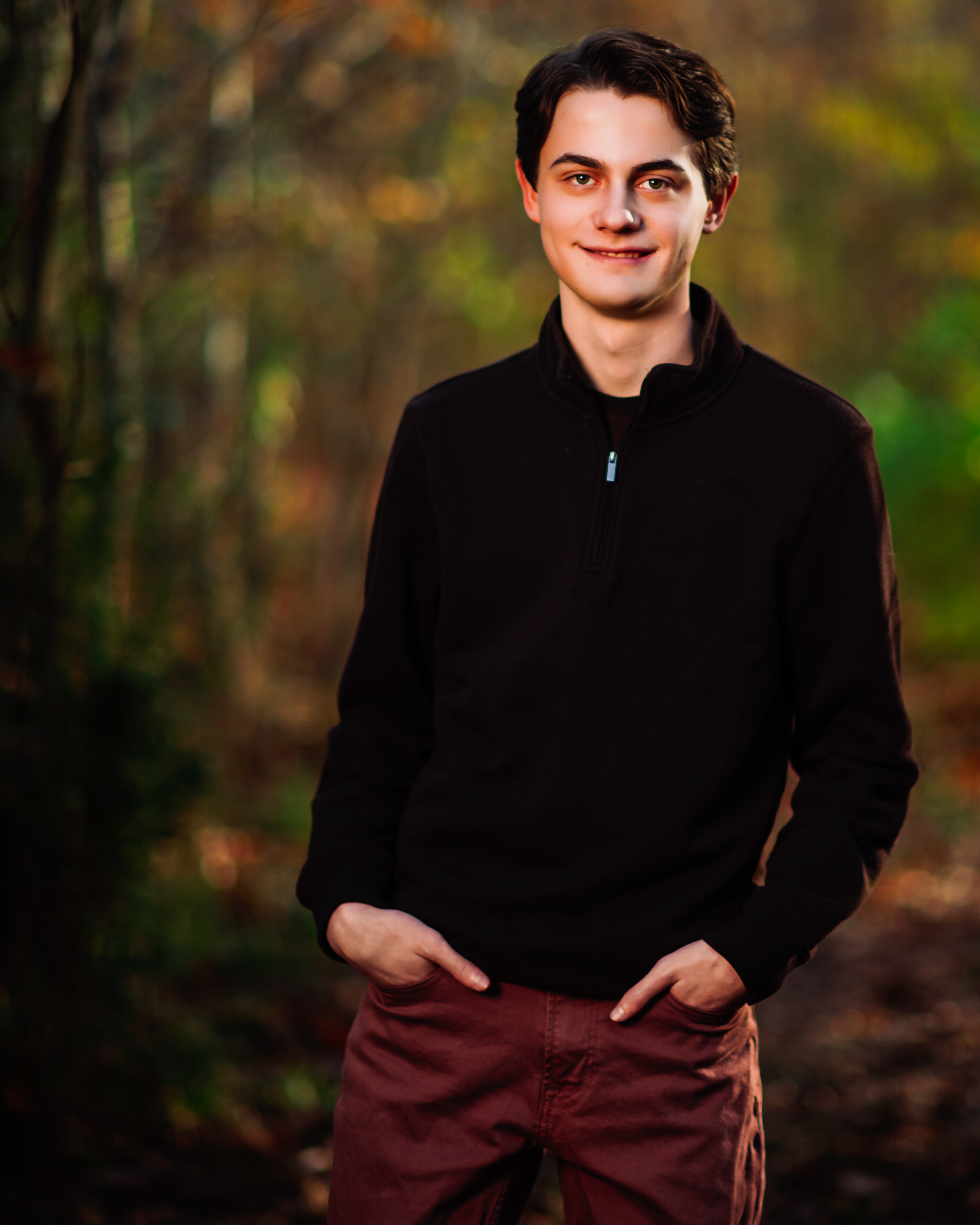 A teenage boy in a dark quarter-zip sweater and rust-colored pants stands easy on a wooded autumn trail, hands in his pockets and a confident half smile, with soft fall color glowing behind him. Fall teen portrait by Nashville family photographer Cate Connery Bury.