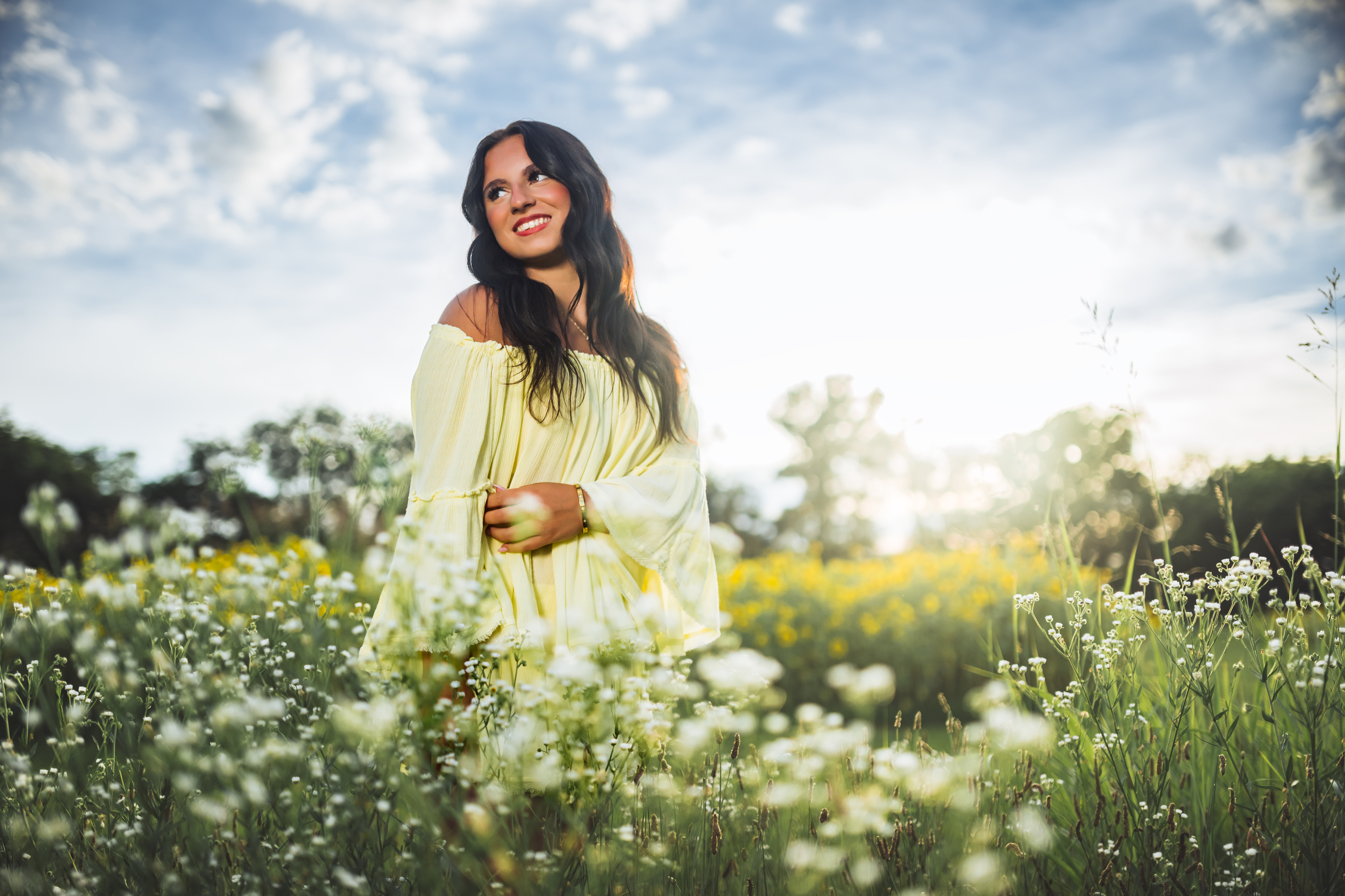 A high school senior in a pale yellow off-the-shoulder dress stands waist-deep in a summer meadow of white wildflowers and golden blooms, smiling as she looks toward the sun breaking through the clouds. A bright, joyful Nashville senior portrait by Cate Connery Bury Photography that feels like the very best day of summer. A high school senior in a pale yellow off-the-shoulder dress stands waist-deep in a summer meadow of white wildflowers and golden blooms, smiling as she looks toward the sun breaking through the clouds. A bright, joyful Nashville senior portrait by Cate Connery Bury Photography that feels like the very best day of summer.