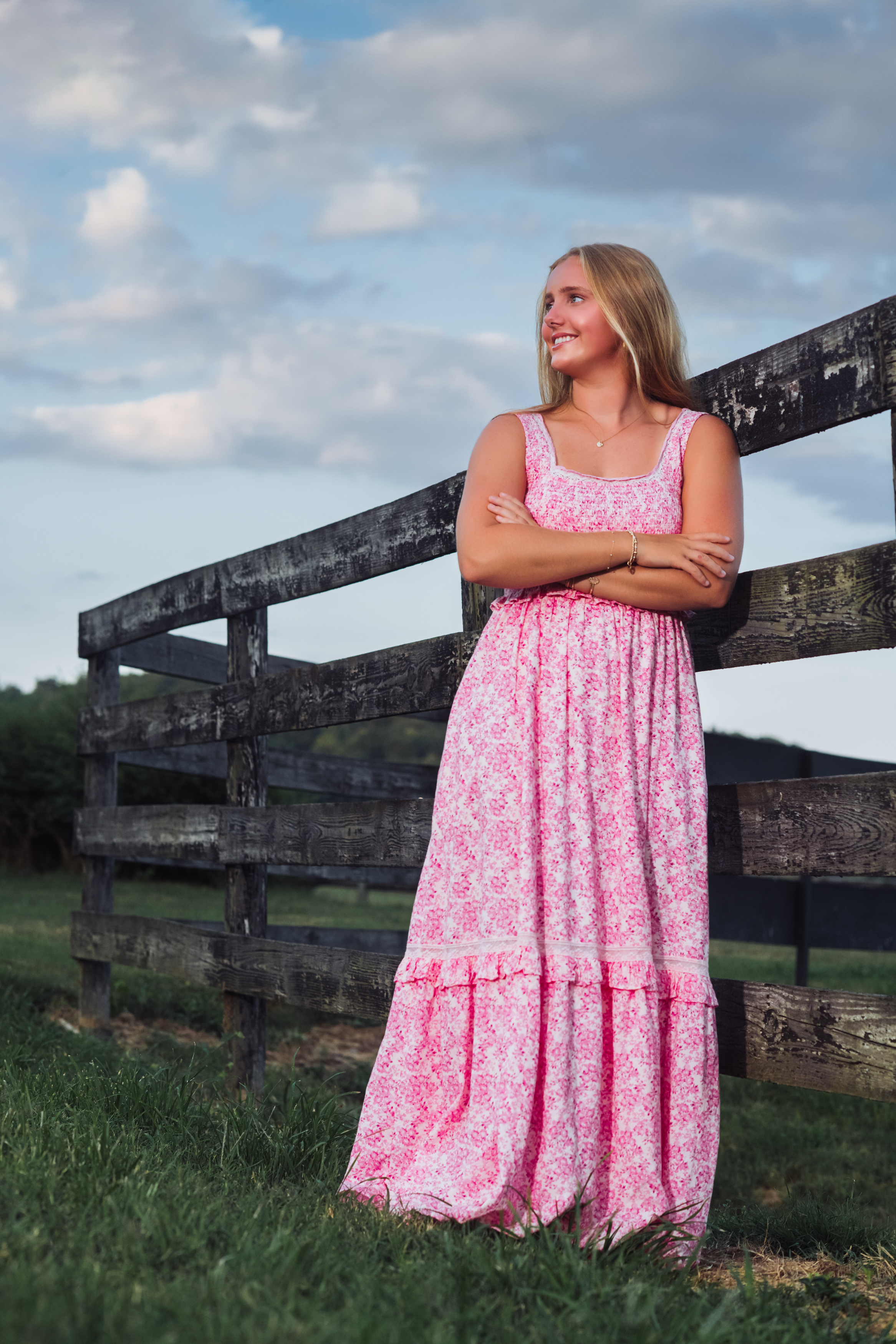 A high school senior in a pink floral smocked maxi dress leans against a weathered wooden fence at the Steeplechase in Warner Parks, arms crossed and smiling as she gazes across the rolling Tennessee hills under a sky full of soft summer clouds. A confident, classic Nashville senior portrait by Cate Connery Bury Photography with true Tennessee countryside charm. A high school senior in a pink floral smocked maxi dress leans against a weathered wooden fence at the Steeplechase in Warner Parks, arms crossed and smiling as she gazes across the rolling Tennessee hills under a sky full of soft summer clouds. A confident, classic Nashville senior portrait by Cate Connery Bury Photography with true Tennessee countryside charm.