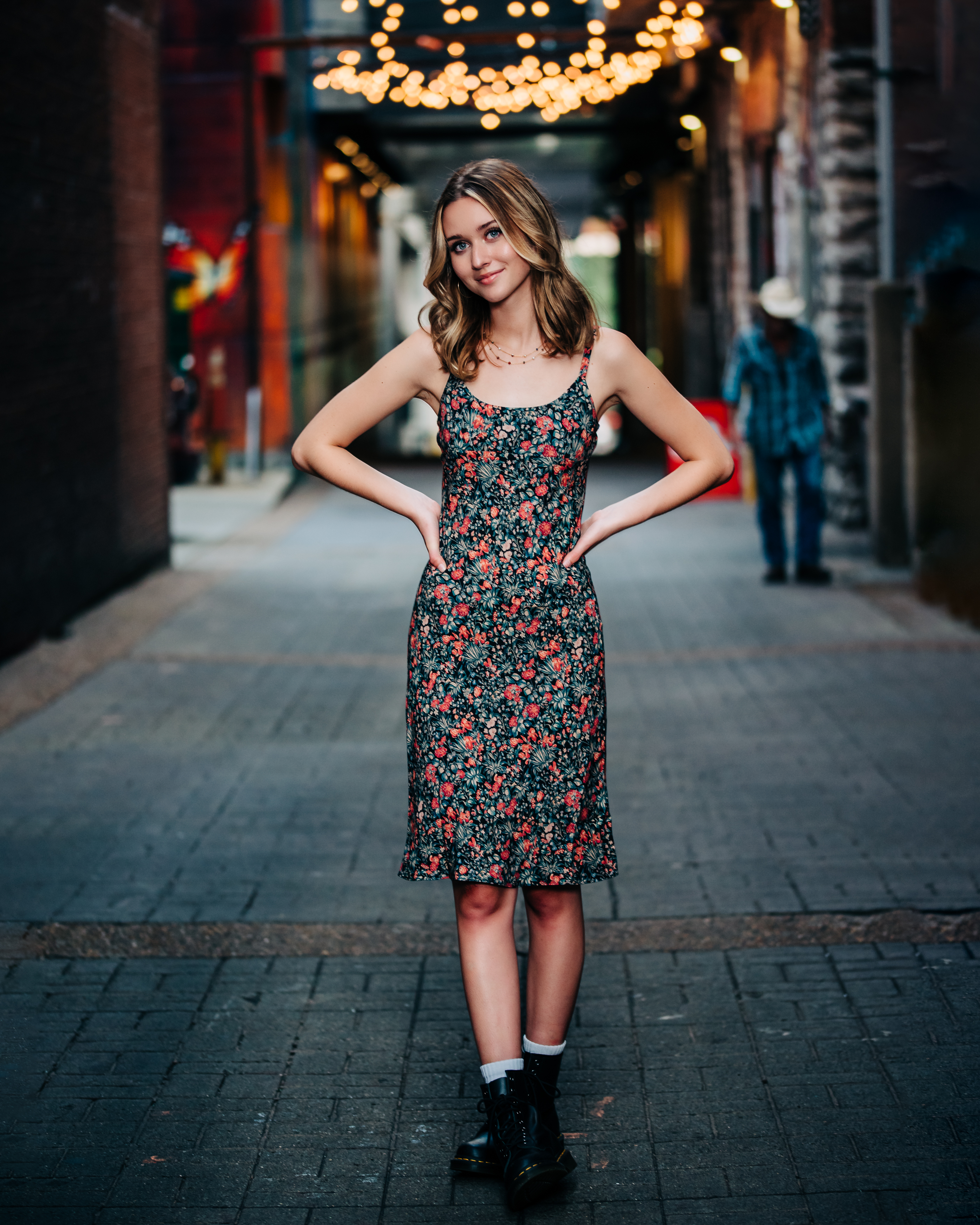 A high school senior in a dark floral slip dress, white socks, and black combat boots stands with her hands on her hips in Printers Alley, head tilted with a playful smile, as glowing string lights, brick walls, and colorful murals stretch out behind her. A spirited, editorial downtown Nashville senior portrait by Cate Connery Bury Photography with true Music City personality. A high school senior in a dark floral slip dress, white socks, and black combat boots stands with her hands on her hips in Printers Alley, head tilted with a playful smile, as glowing string lights, brick walls, and colorful murals stretch out behind her. A spirited, editorial downtown Nashville senior portrait by Cate Connery Bury Photography with true Music City personality.