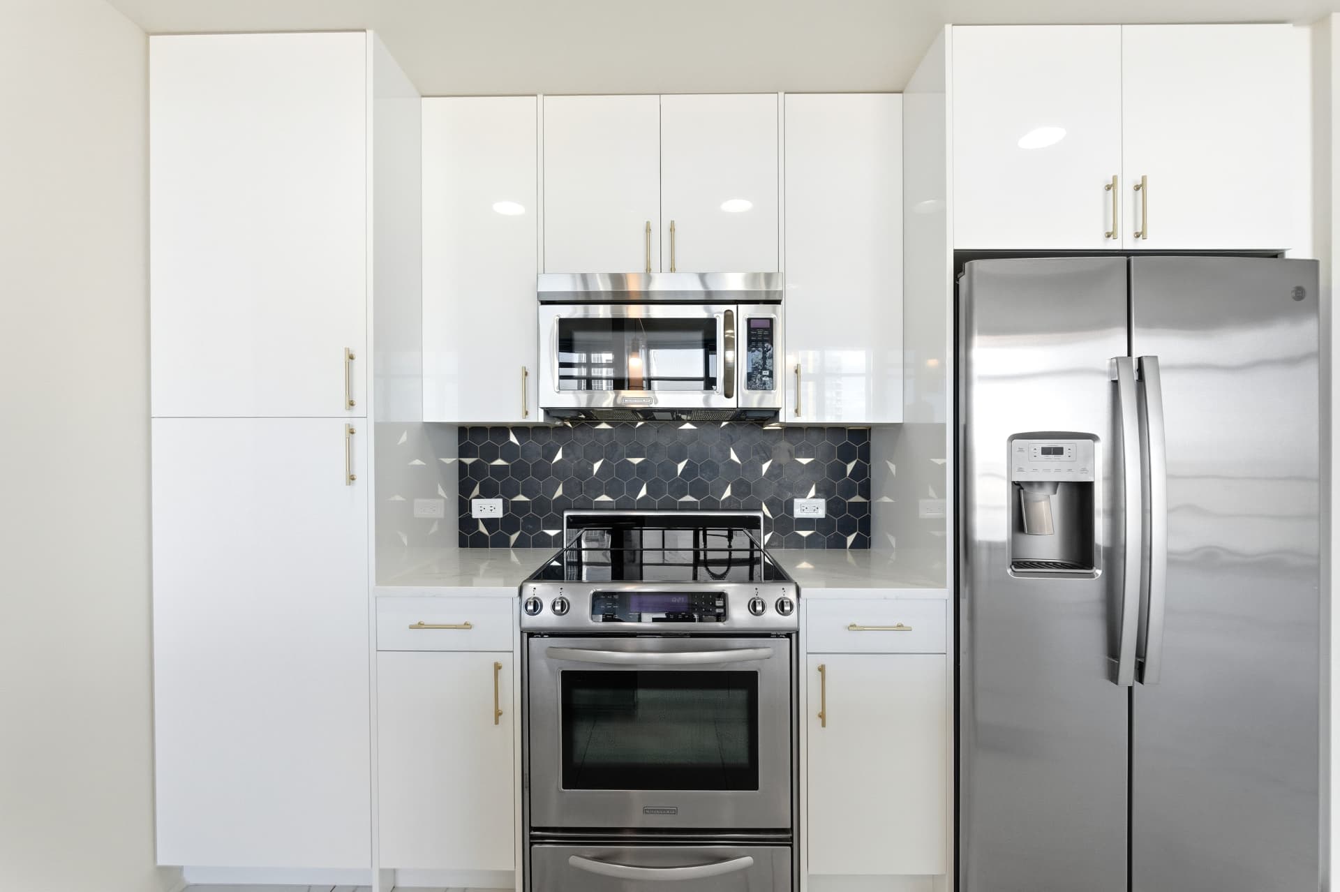 Kitchen cook wall with stainless range and microwave, charcoal hex tile backsplash and high-gloss white cabinetry with gold pulls