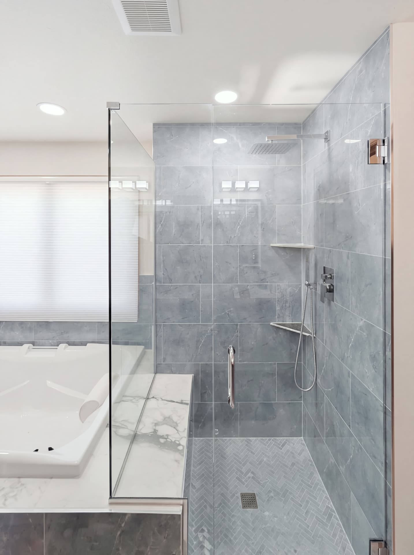 Walk-in shower with blue-grey herringbone floor tile, chrome rainfall showerhead and frameless glass door