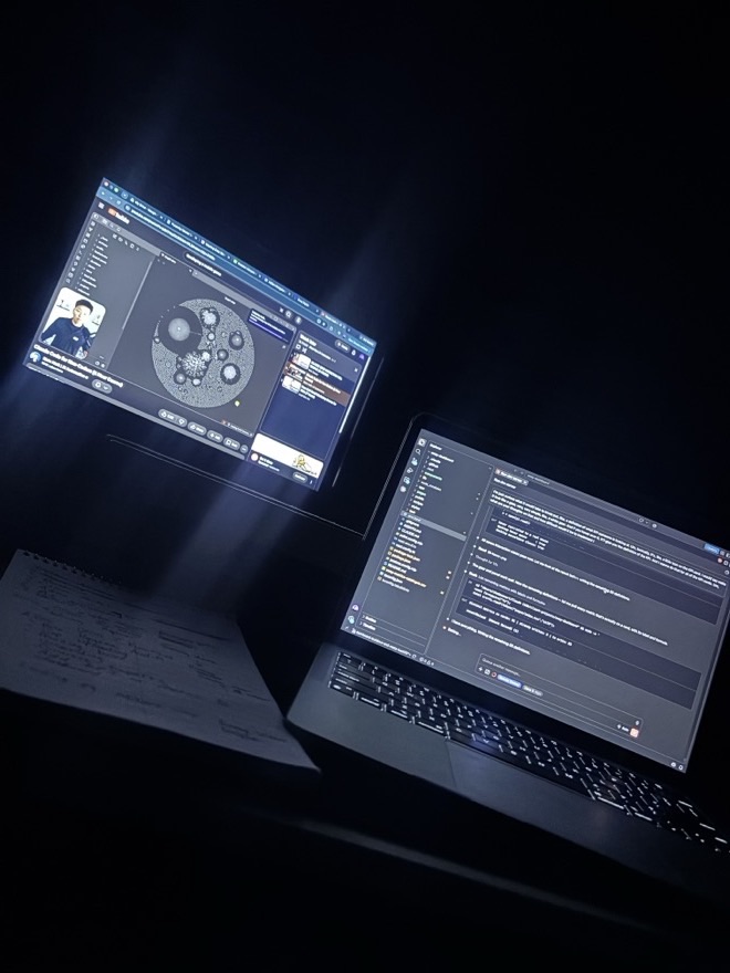 Late-night desk setup: a business course playing on one screen, code on the laptop, handwritten notes beside it