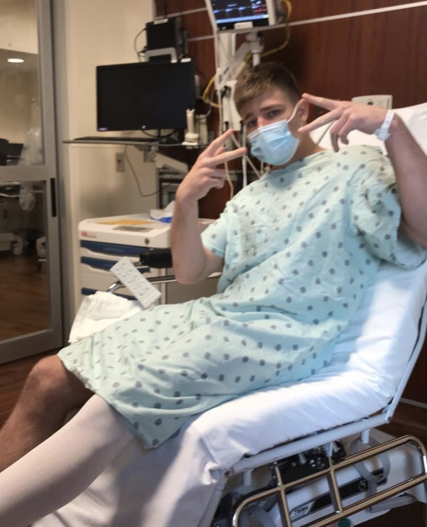 Chad in a hospital gown after ACL surgery, throwing peace signs