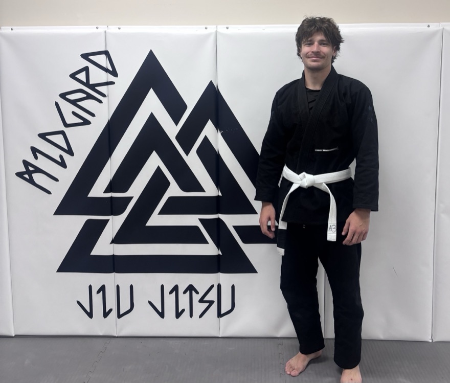 Chad in a black gi and white belt in front of the Midgard Jiu-Jitsu logo