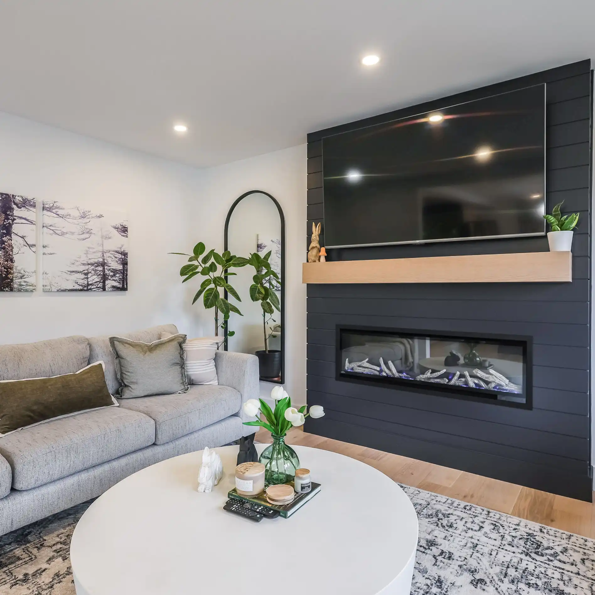 Modern living room with gray sofa, round table, TV above a fireplace, and plants. Art on wall, rug on floor, cozy atmosphere.