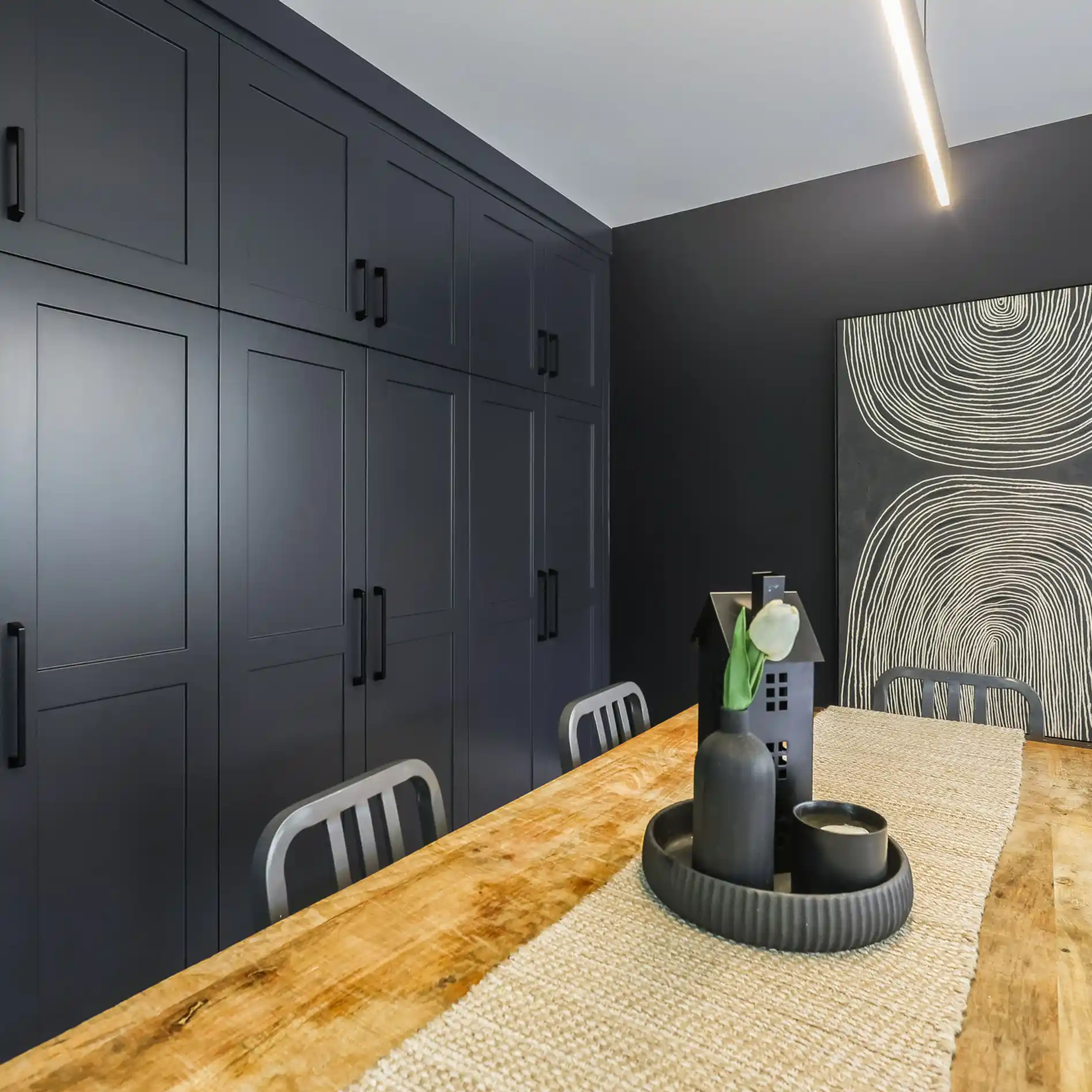 Modern dining room with dark cabinets, abstract art, and a wooden table. A black vase with a tulip on a woven runner adds elegance.