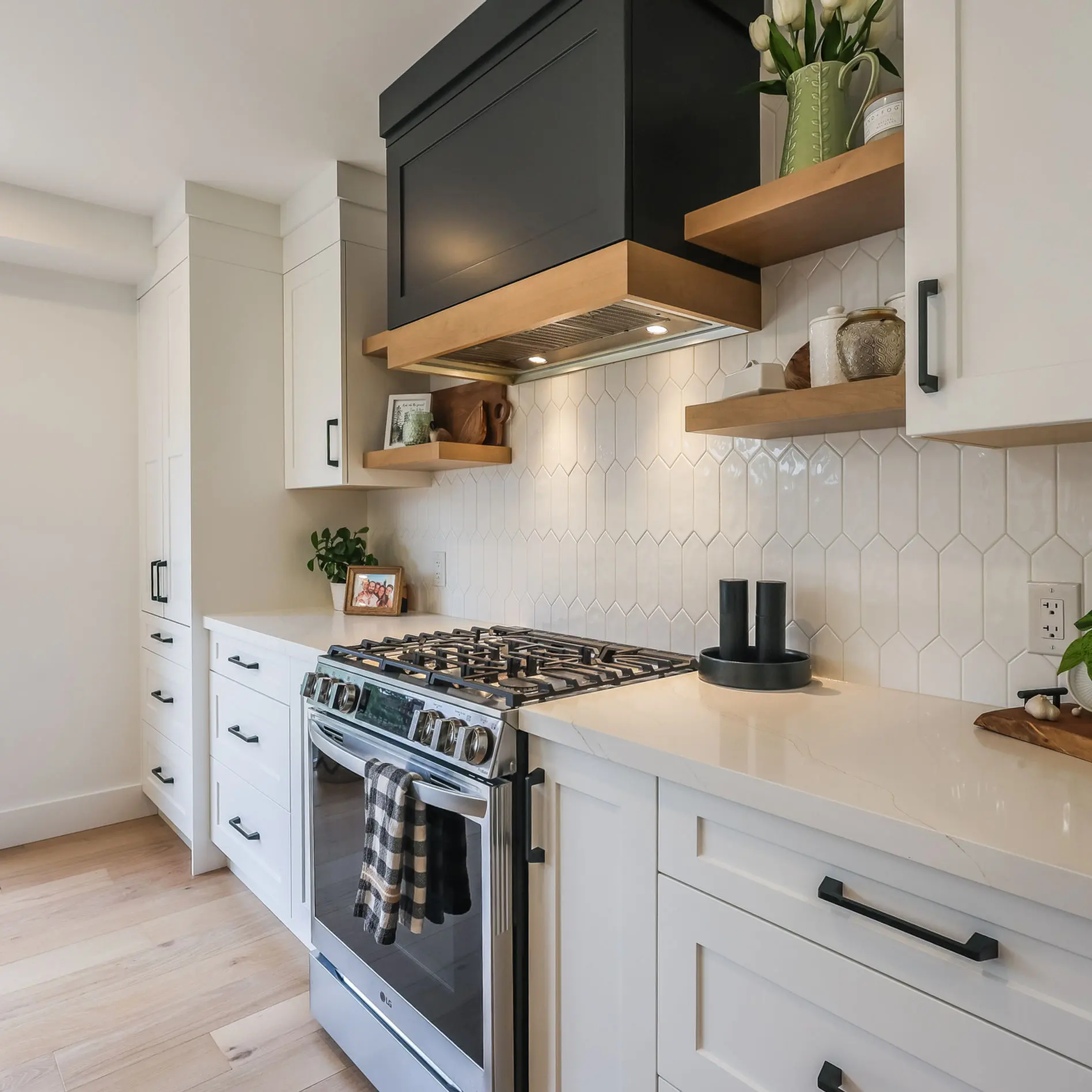 Modern kitchen with white cabinets, black hood, plants, and a gas stove. Hexagon-tiled backsplash. Bright light and greenery outside.