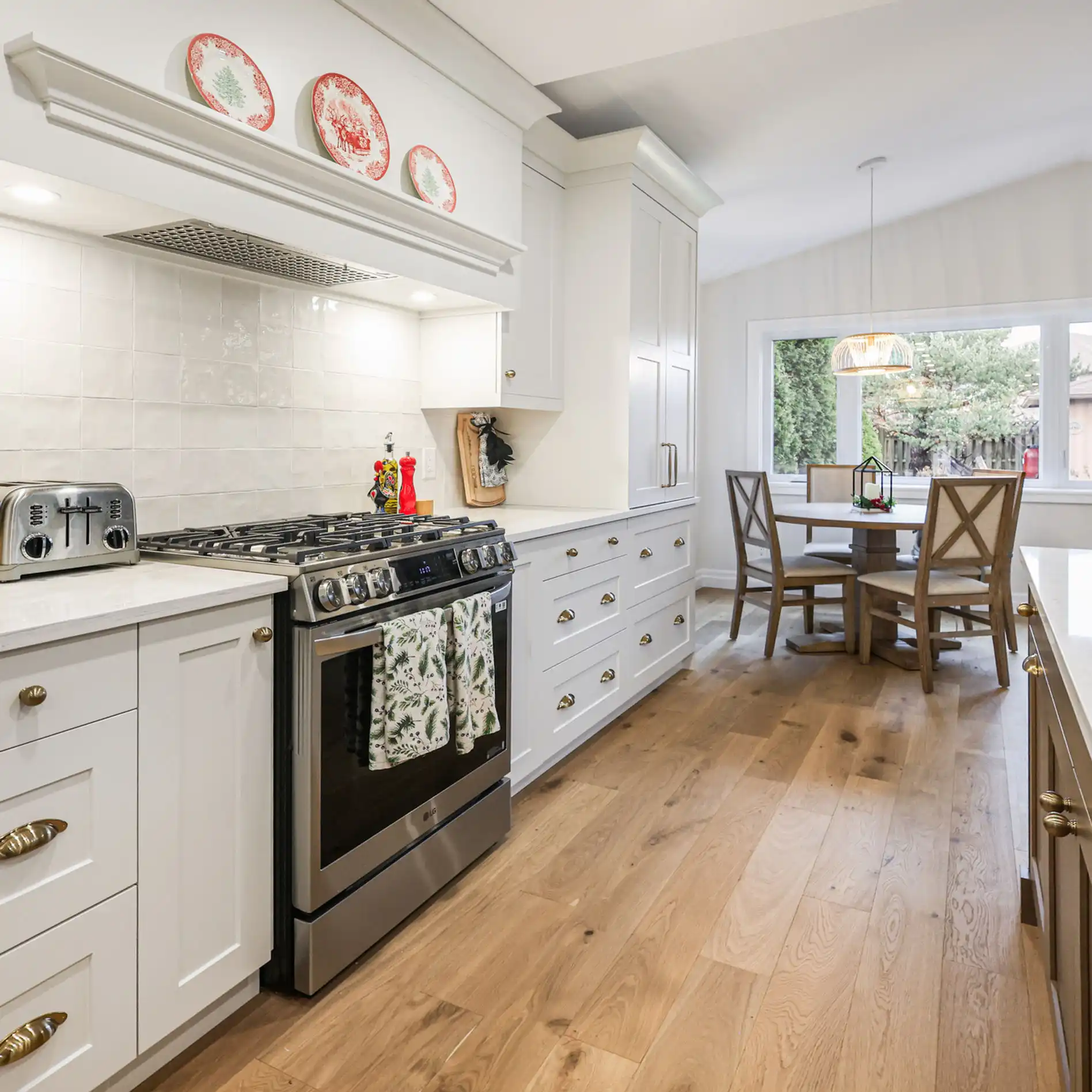 Modern kitchen with white cabinets, stove, and decorative plates. Wooden table and chairs by large window, light wood floor, cozy atmosphere.