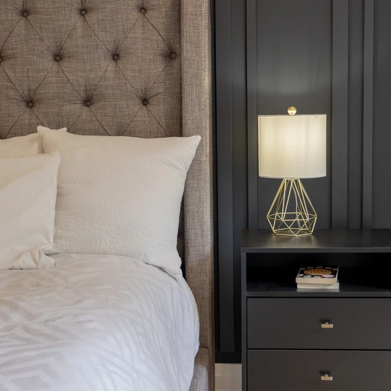 Bed with white pillows and patterned duvet, beside a nightstand with a book and a lamp with gold wireframe base, against a dark wall.