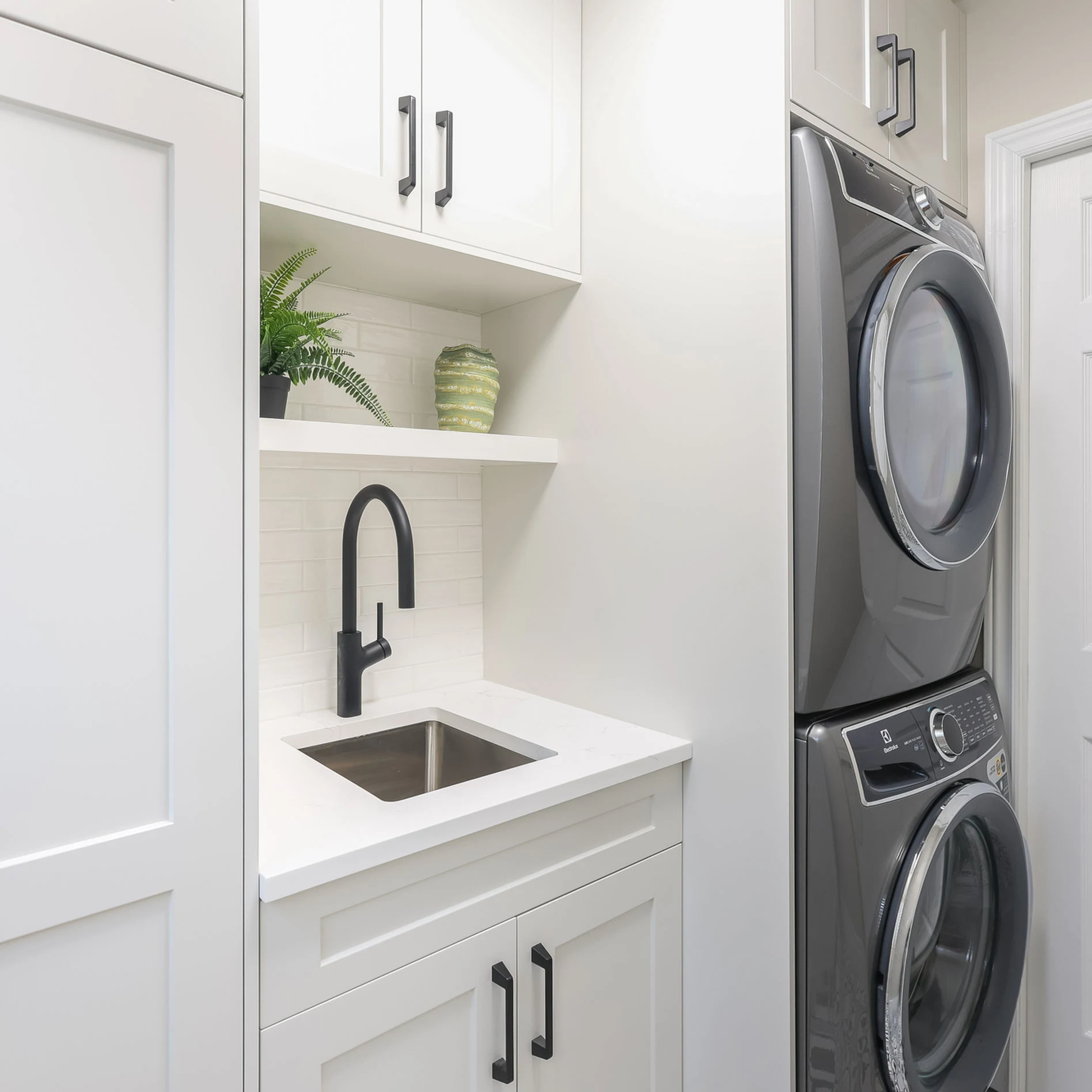 White laundry room with stacked grey washer-dryer, black faucet sink, and green plant on shelf. Clean, modern design.