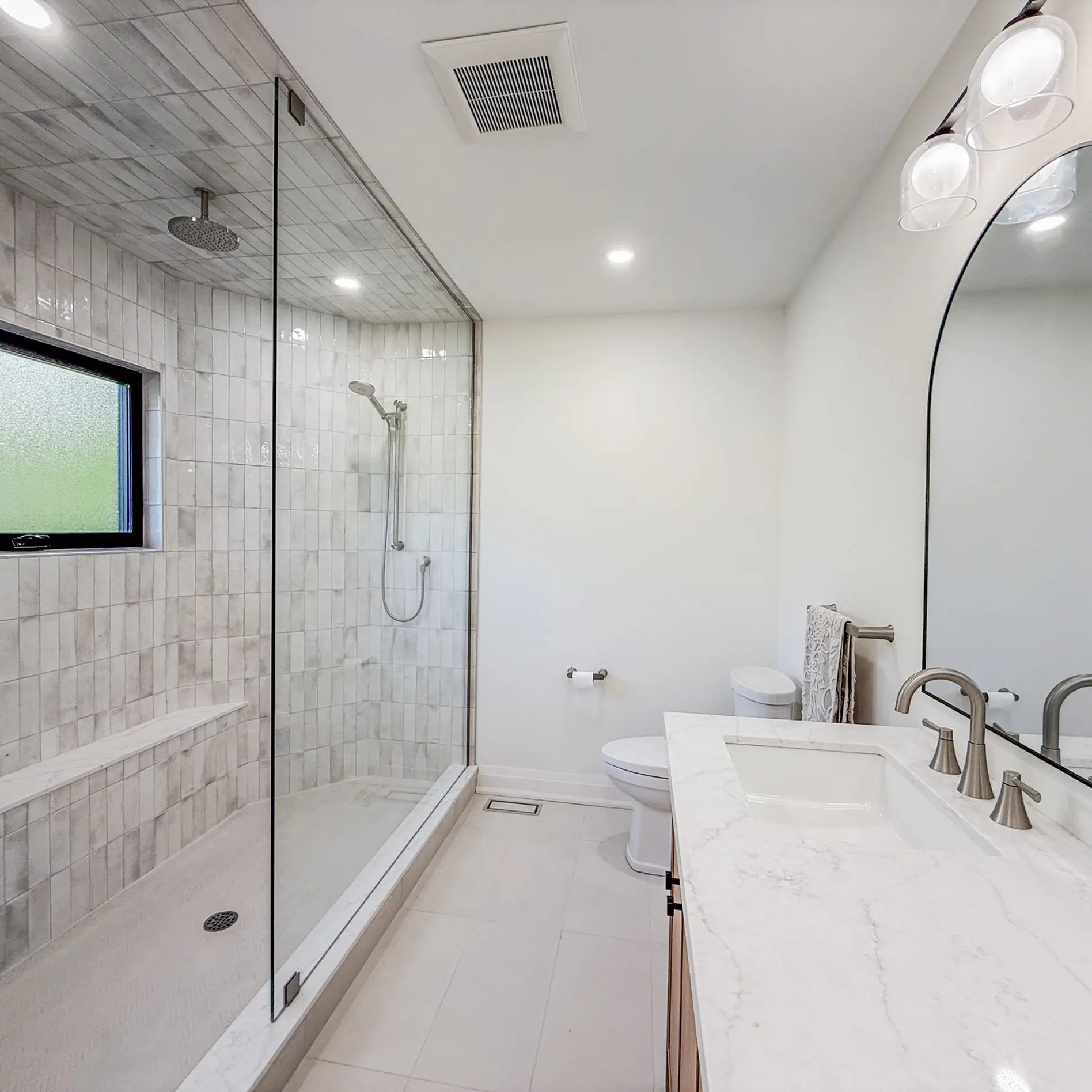 Modern bathroom with glass shower, marble tiles, white countertop sink, oval mirror, and silver fixtures. Bright and minimalistic design.
