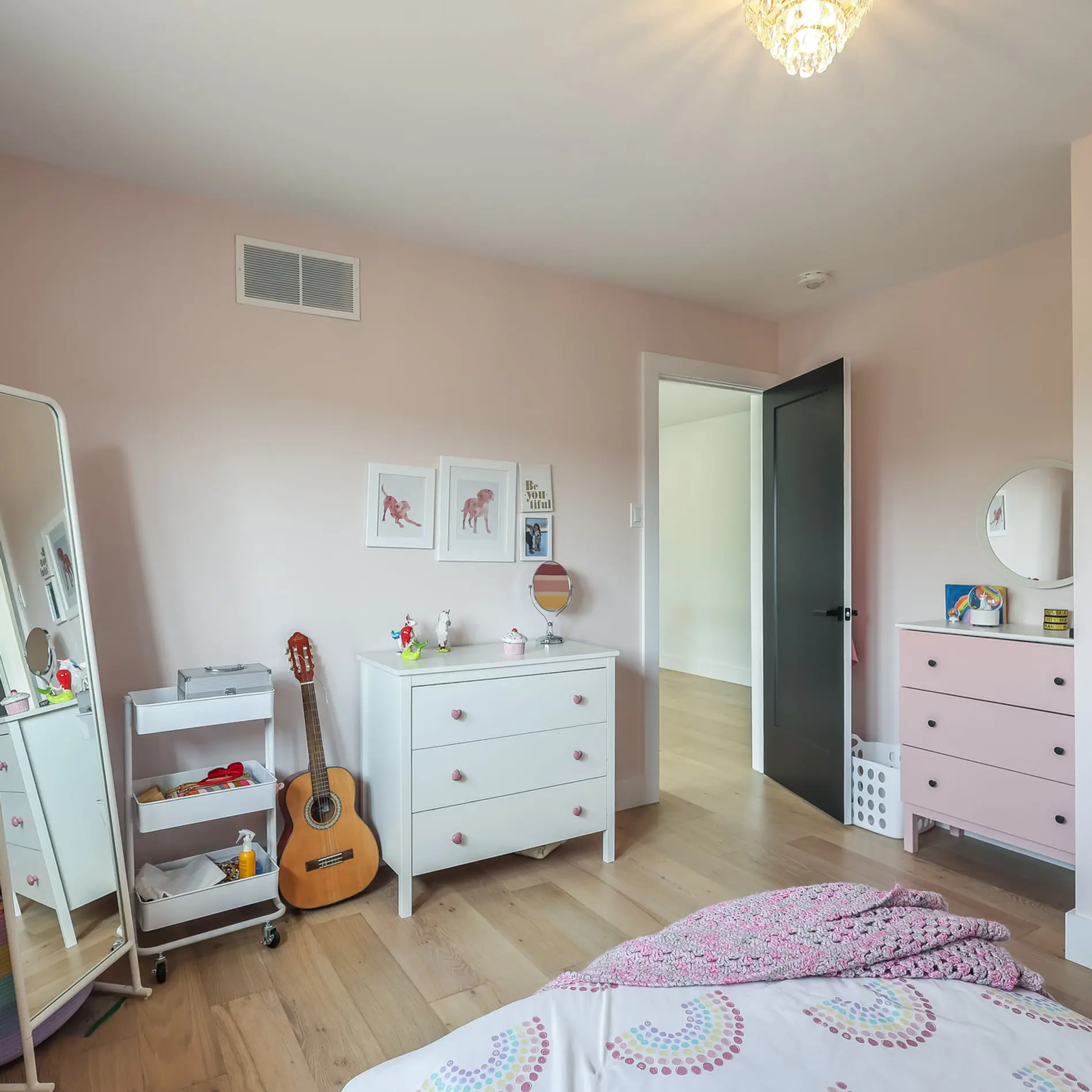 A pink-walled bedroom with a bed, guitar, standing mirror, and dresser. The sunlight filters through a window, creating a cozy atmosphere.