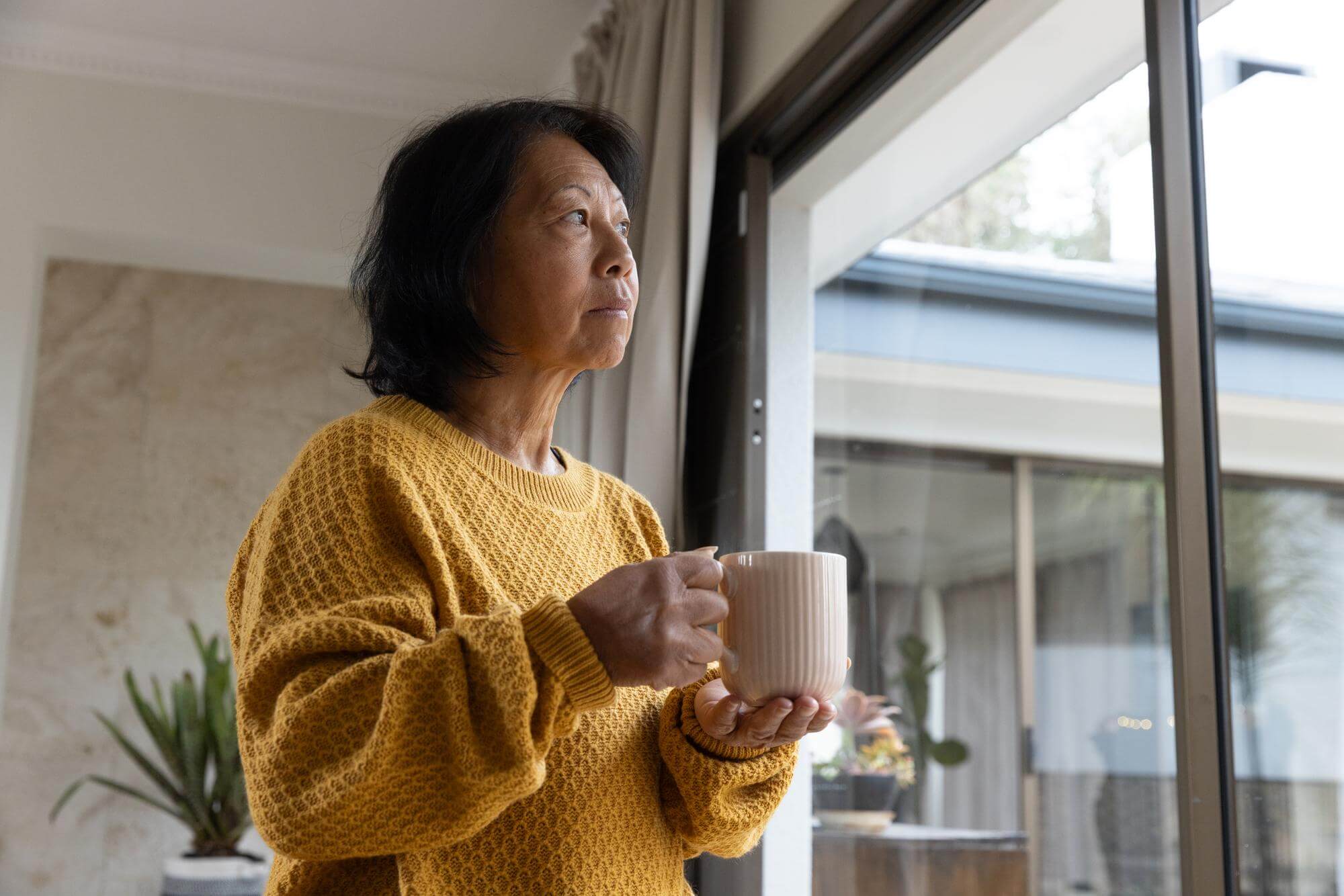 Senior Asian woman holding a mug, gazing out a glass door in morning light