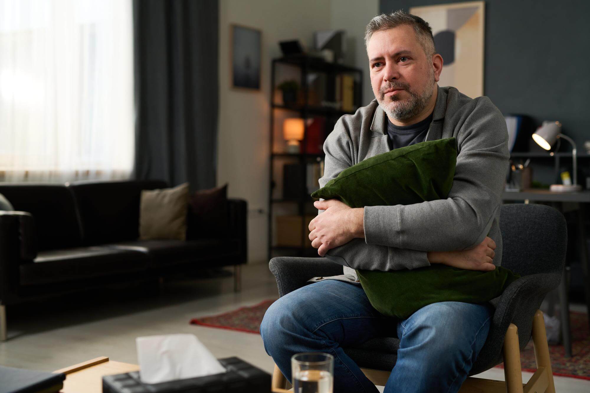 Middle-aged man sitting quietly on a chair hugging a cushion in a muted living room