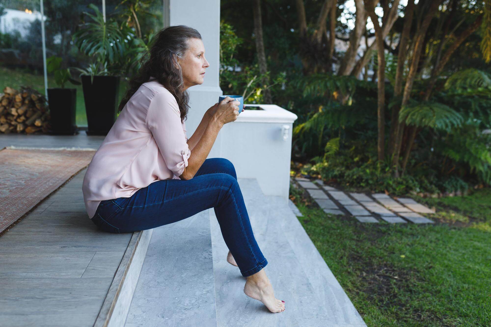 Senior woman sitting barefoot on a garden step with a morning coffee