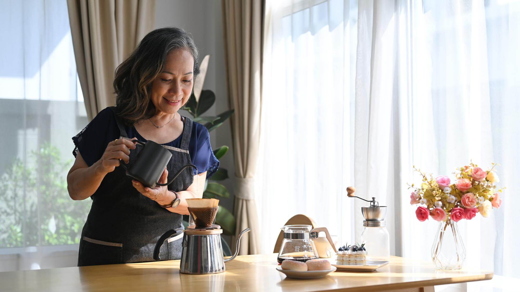 Asian woman in an apron pouring a kettle over a coffee dripper at a sunlit kitchen table