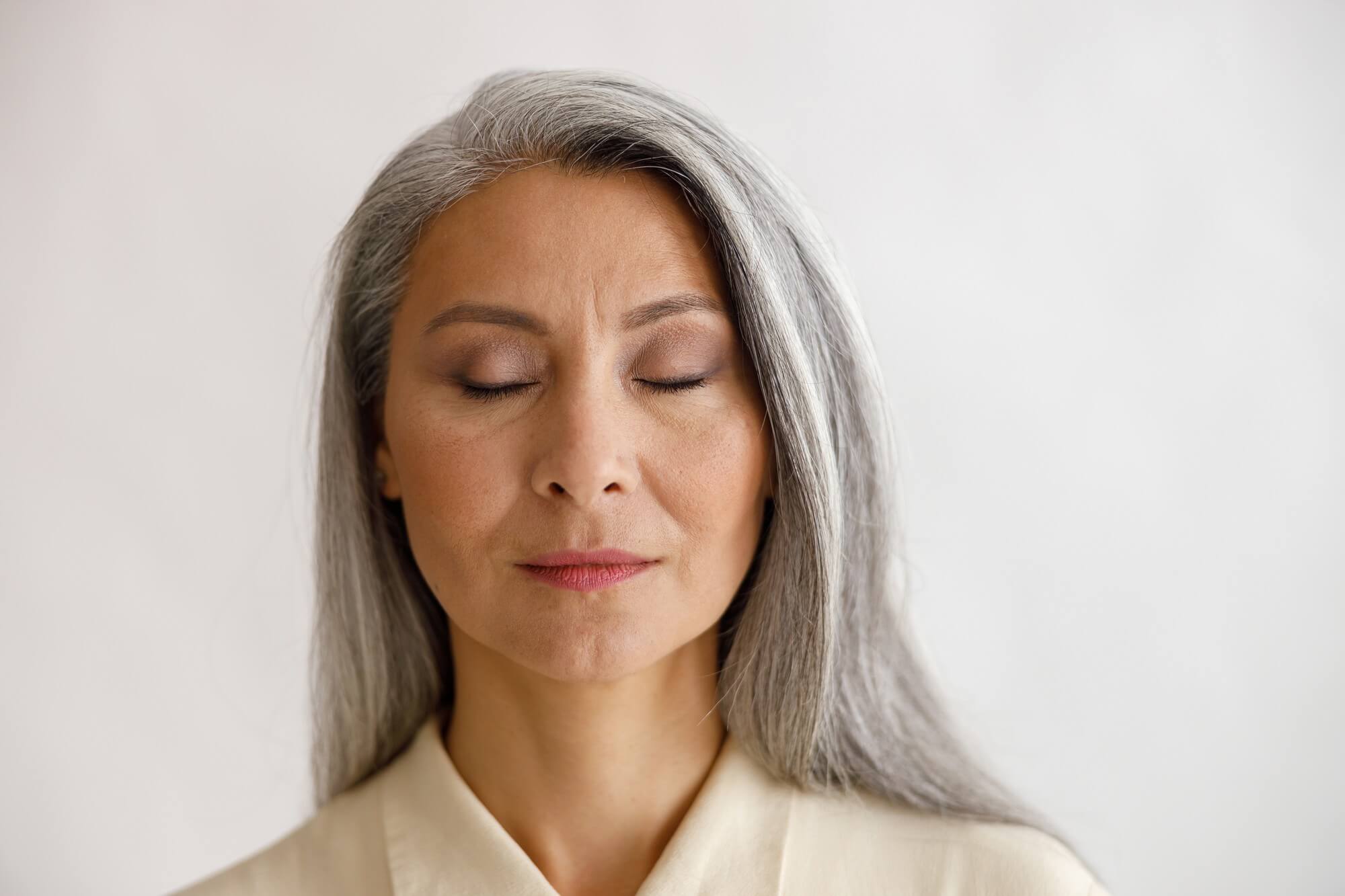 Close portrait of a gray-haired woman with eyes closed against a plain cream background