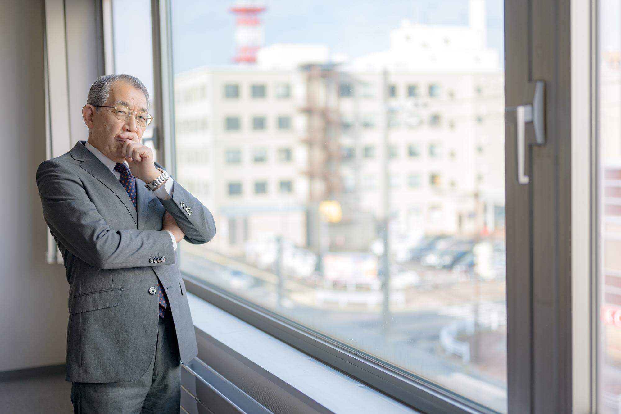 Senior Japanese businessman pausing at an office window, hand to his chin, city outside