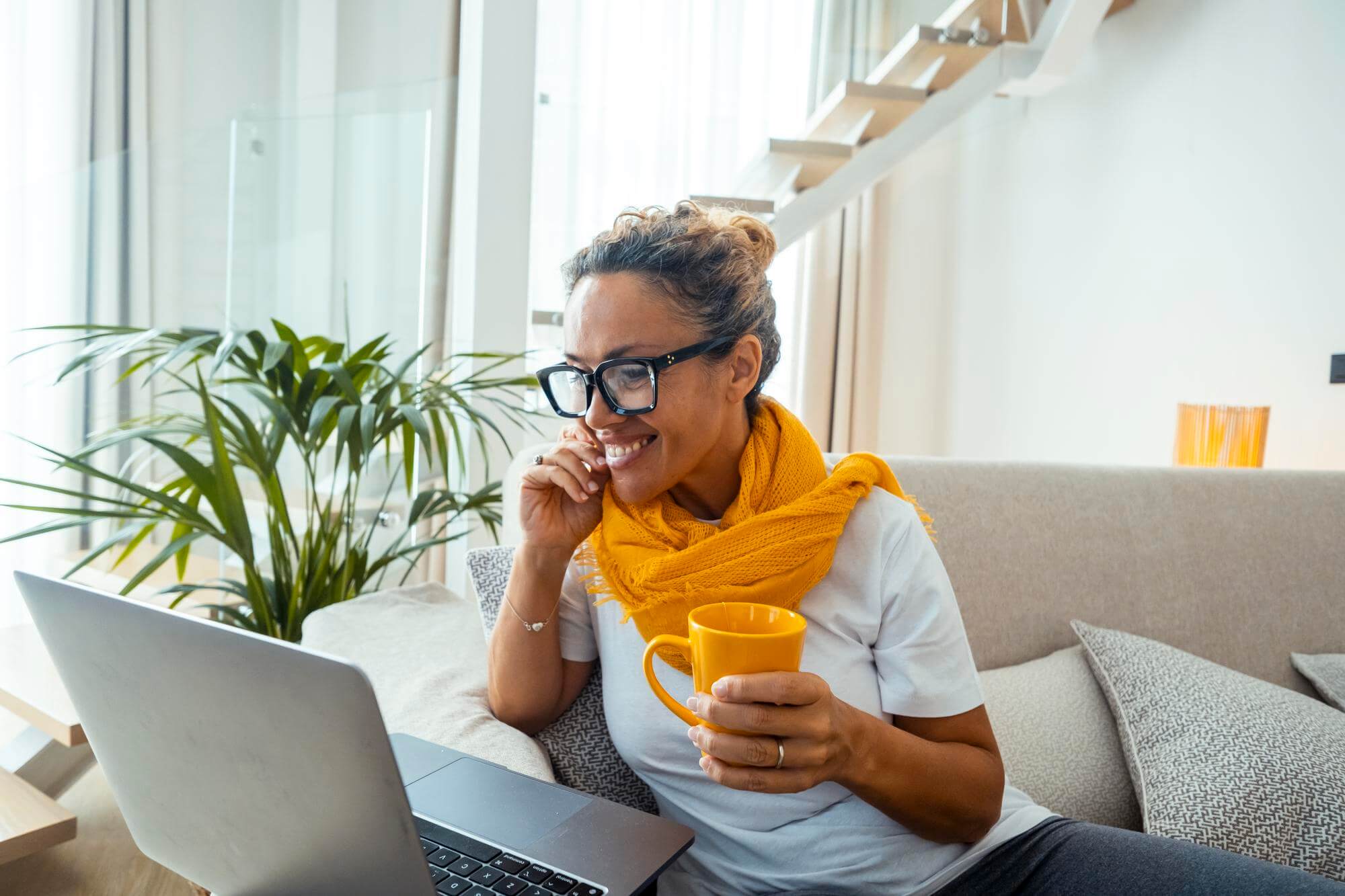 Black woman in glasses and a yellow scarf holding a mug, listening to a laptop call on a sofa