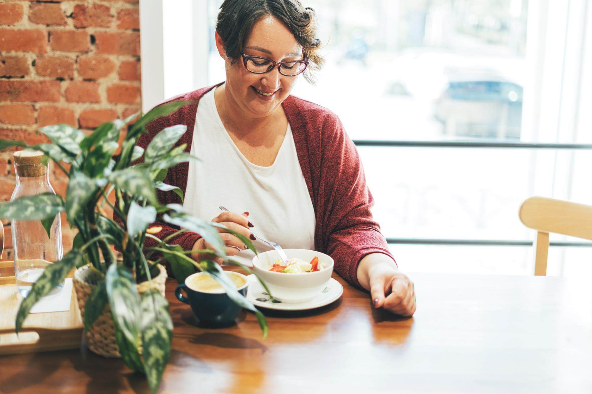 Woman in glasses eating a simple bowl at a sunlit kitchen table with plants