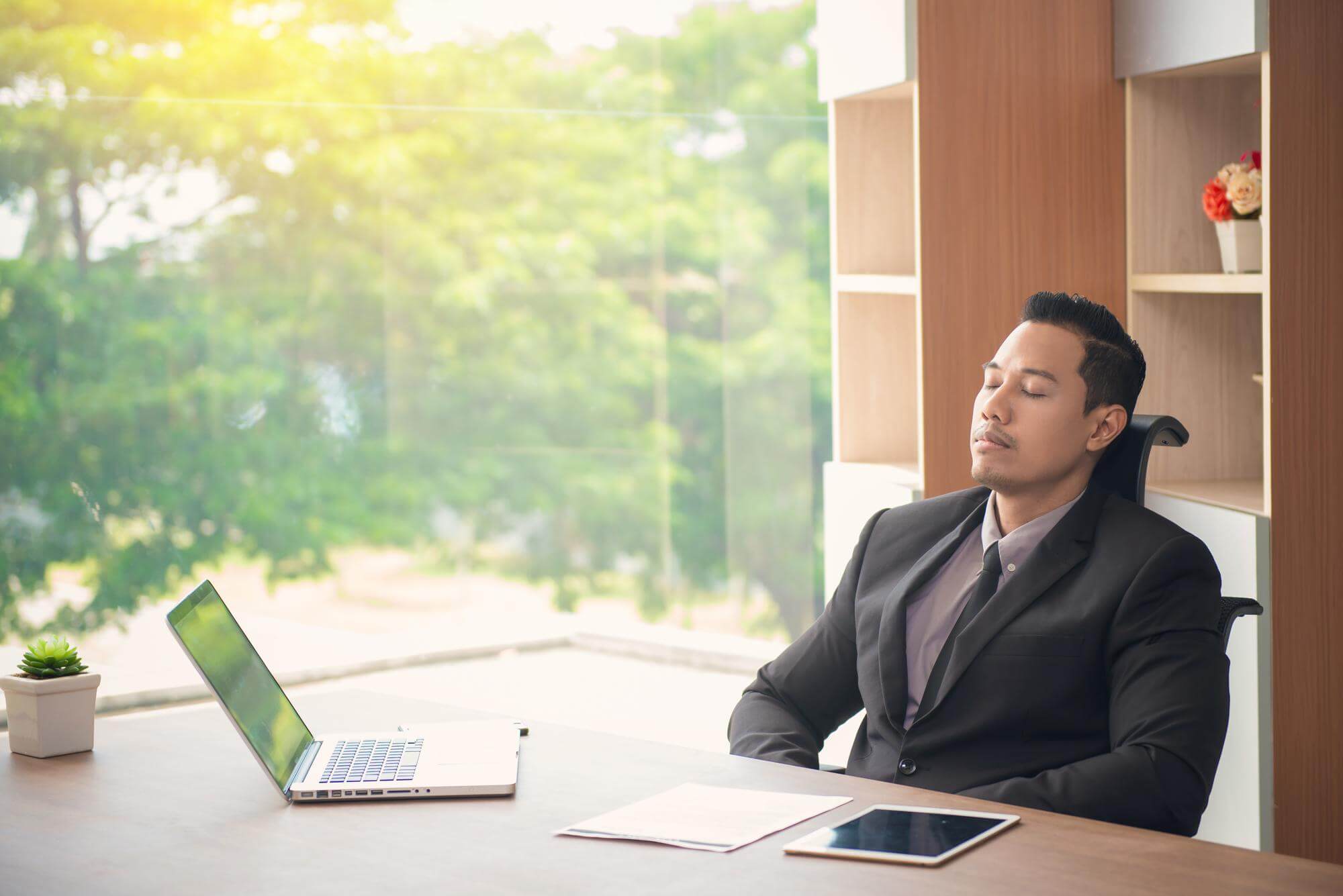 Asian man at a desk by a sunlit window, eyes closed, taking a deliberate breath