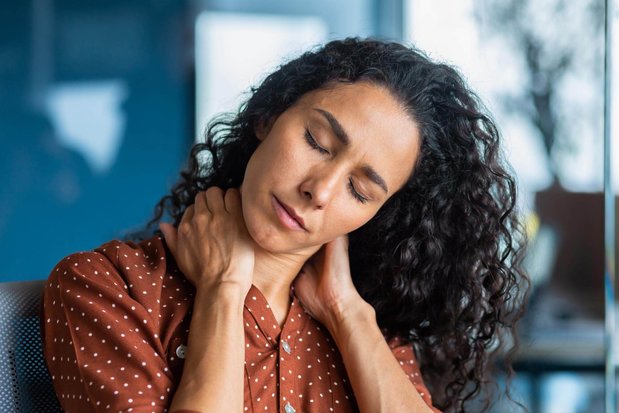 Latina woman with curly hair, eyes closed, hands resting on the back of her neck