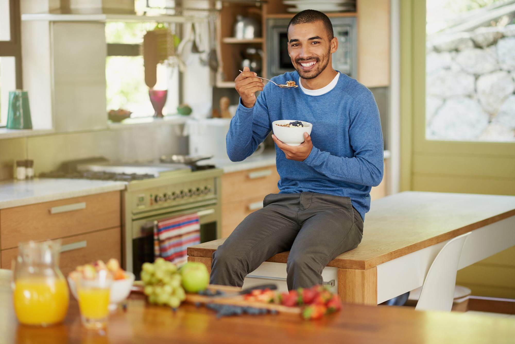Man perched on a kitchen counter eating a bowl of fruit, smiling