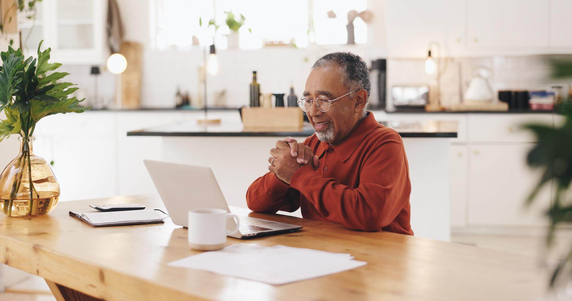 Senior Black man at a kitchen table on a laptop video call, hands clasped, listening calmly