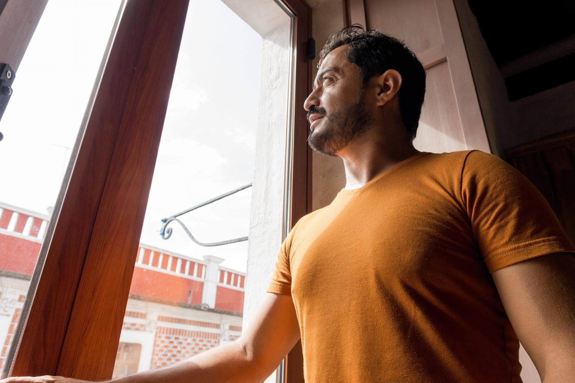 Bearded Hispanic man in an orange shirt looking out an open window, posture easy
