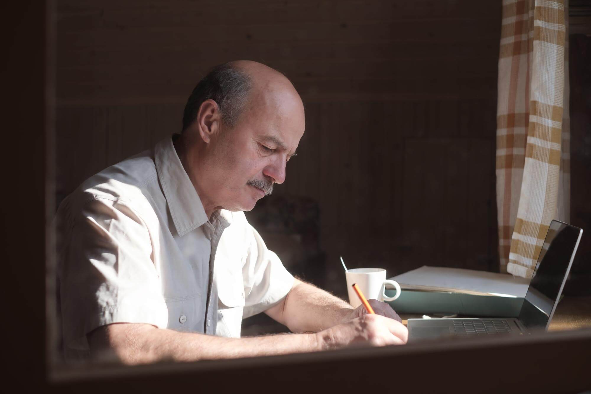Senior Hispanic man writing in a journal at a table beside a window