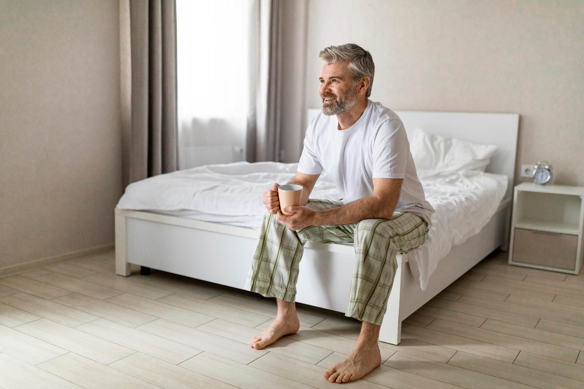 Gray-haired man sitting on the edge of his bed holding a mug in bright morning light, no phone