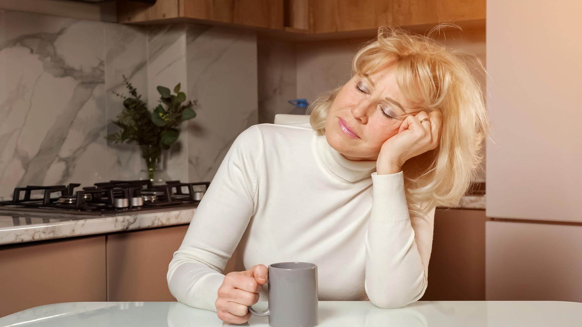 Tired middle-aged woman resting her head on her hand with a coffee at the kitchen counter