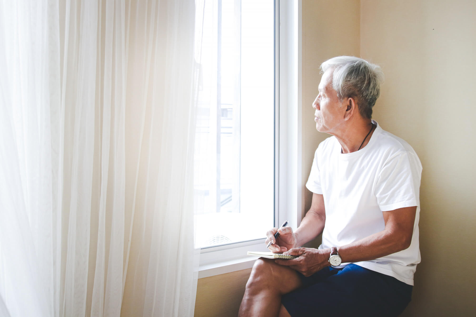 Senior Asian man seated by a bright window in soft light, quietly contemplative