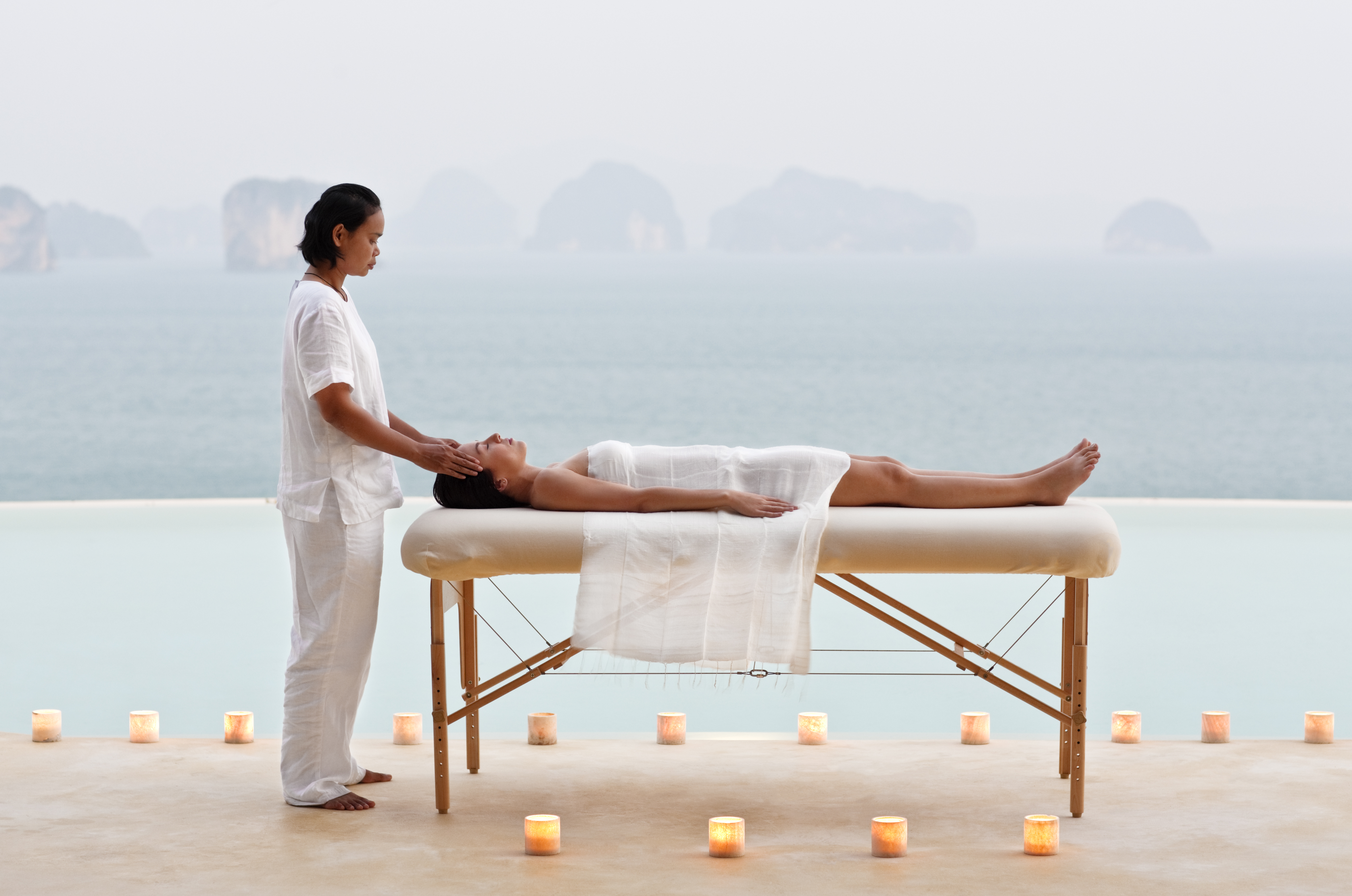 Woman lying peacefully on a massage table as a therapist in white rests her hands on her head, surrounded by glowing candles overlooking misty islands and the sea