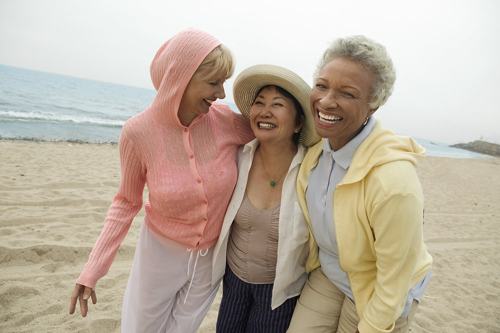 Three smiling older women with arms around each other, laughing together on a sandy beach by the ocean