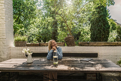 Woman sitting at a rustic wooden table in a sunlit garden, eyes closed and smiling, with a jug of white flowers and a pitcher of lemon water