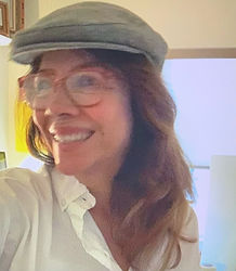 Smiling woman with long auburn hair wearing glasses, a grey flat cap and a white shirt