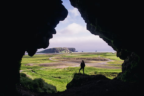 Silhouette of a person standing at the mouth of a dark cave, looking out over green fields, a winding path and a rocky sea stack under a bright sky