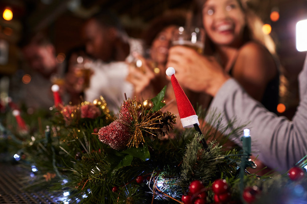 Friends laughing and toasting with drinks at a festive holiday party, with pine garland, red berries and a tiny Santa hat in the foreground