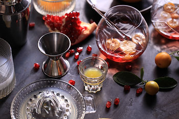 Mocktail-making setup on a dark table with a glass mixing pitcher, a jigger, a citrus juicer, fresh pomegranate seeds and kumquats Mocktail-making setup on a dark table with a glass mixing pitcher, a jigger, a citrus juicer, fresh pomegranate seeds and kumquats