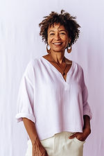 Smiling woman with short curly hair in a soft lavender blouse, standing with her hands in her pockets against a pale backdrop