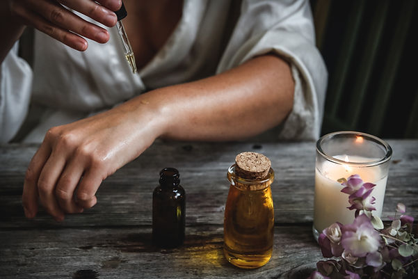 Woman applying essential oil to her wrist with a dropper, beside amber oil bottles, a lit candle and dried purple flowers on a rustic wooden table