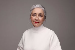 Elegant older woman with silver hair swept back, red lipstick and a white turtleneck sweater, smiling softly against a grey backdrop