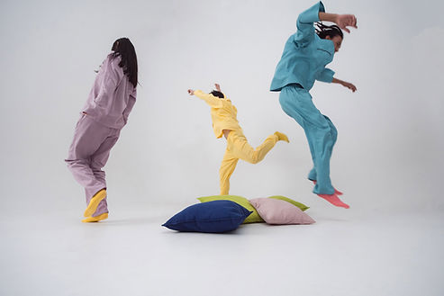 Three people in colorful pastel pajamas leaping and dancing playfully over a pile of cushions against a white backdrop Three people in colorful pastel pajamas leaping and dancing playfully over a pile of cushions against a white backdrop